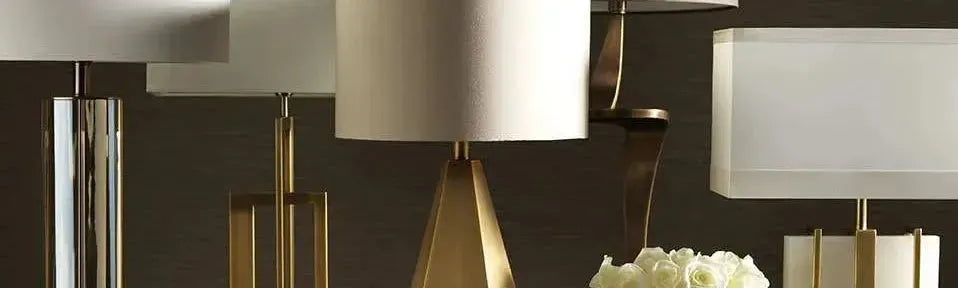 Assorted brass and metallic table lamps with white fabric shades arranged against a dark textured wall background