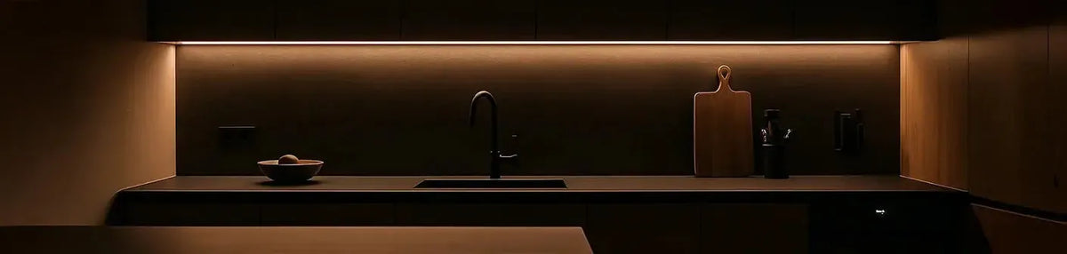 Warm LED strip lighting mounted under dark timber cabinetry above a modern matte black kitchen sink and benchtop