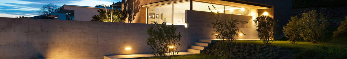 Warm wall lights mounted on a concrete exterior wall illuminating a modern garden path at dusk