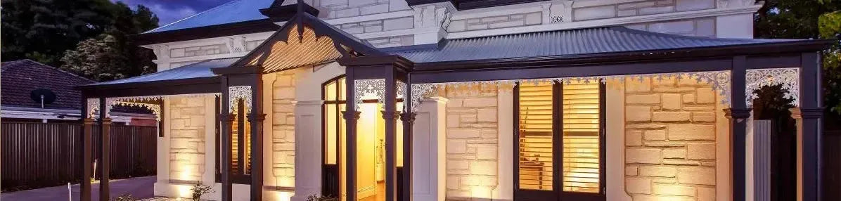 Warm wall lights illuminate the stone facade and ornate white fretwork of a traditional Federation style home exterior
