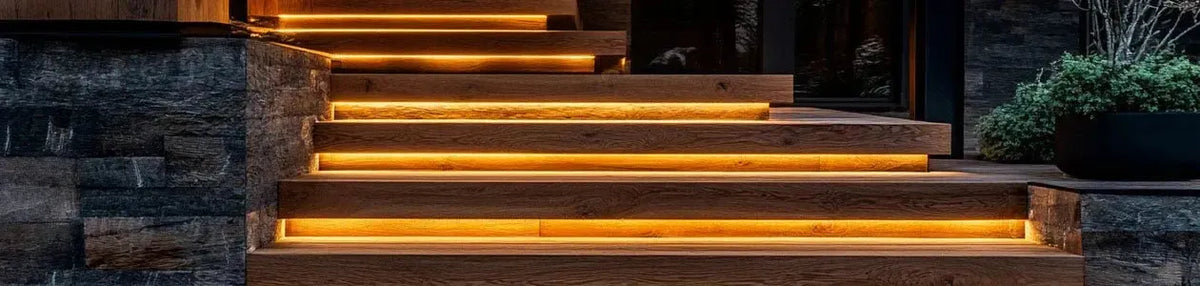 Warm LED strips recessed under timber stair treads against a dark stone wall in an outdoor garden setting