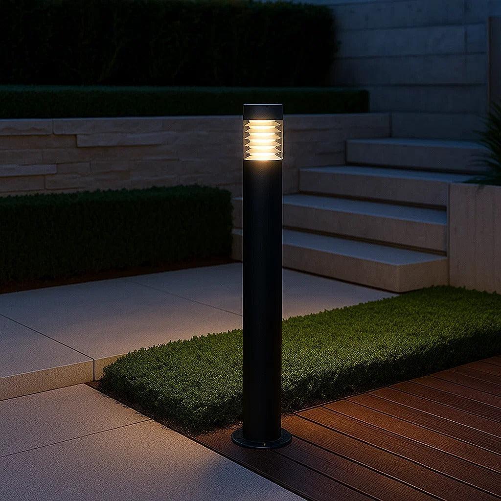 Outdoor black cylindrical bollard light with ribbed diffuser illuminating a modern pathway.