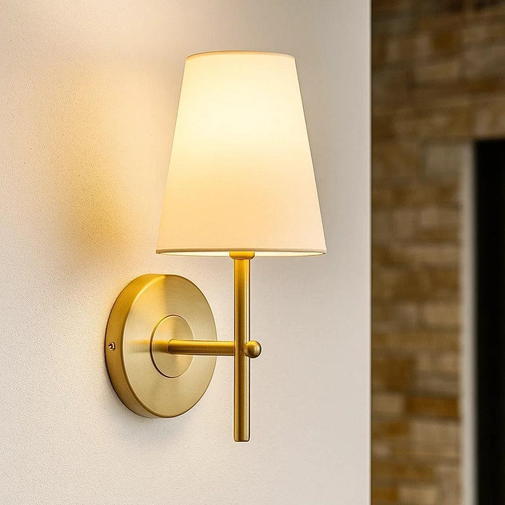 Elegant wall lamp with a brushed brass finish and a tapered white linen shade mounted on a white interior wall.
