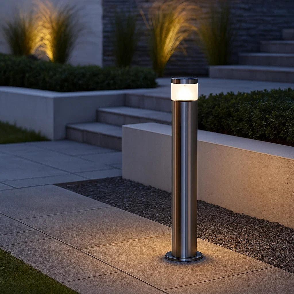 Outdoor stainless steel bollard light illuminating a modern garden path at dusk.