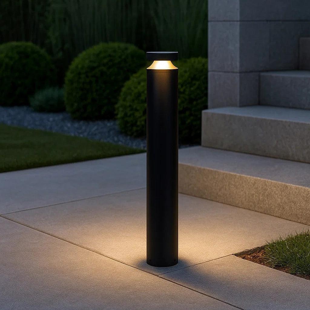 Outdoor black cylindrical bollard light illuminating a concrete path at dusk.