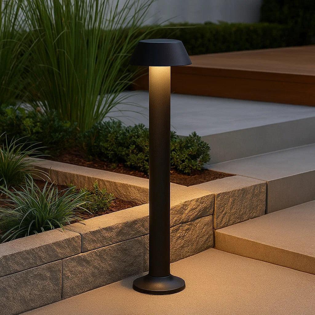 Outdoor black modern bollard light illuminating a concrete pathway and garden.